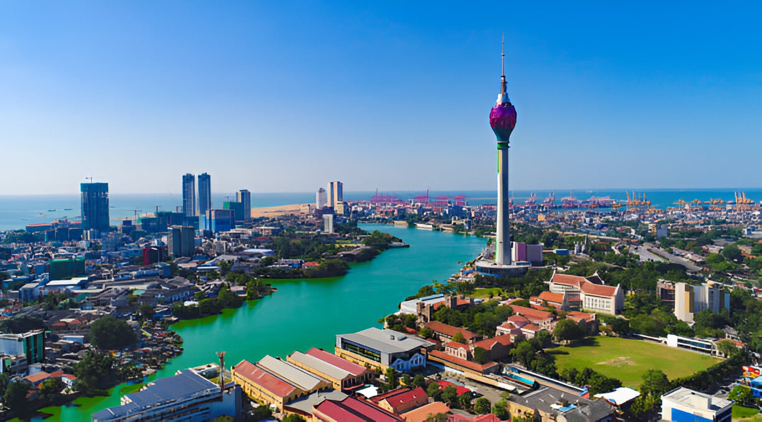 This image showcases a vibrant cityscape of Colombo, Sri Lanka, with the iconic Lotus Tower prominently featured. The tower, with its unique lotus flower design at the top, stands tall against a backdrop of modern skyscrapers and the expansive Indian Ocean. The Beira Lake, with its striking green waters, winds through the city, adding to the scenic beauty. The bustling port in the background highlights Colombo's significance as a major maritime hub. The image captures a clear, sunny day, emphasizing the city's dynamic blend of modernity and natural beauty.